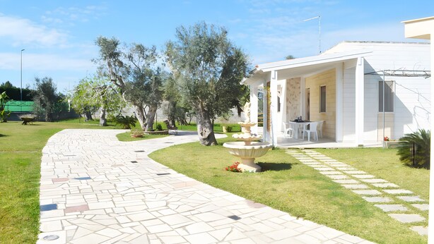 A pathway leading to a white building with a covered patio area, surrounded by greenery and a stone-tiled walkway.