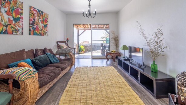 A living room with a brown sofa, colorful cushions, a woven rug, a TV on a black stand, and a green vase with branches. - Country House 'Serrão' with Private Terrace, Private Garden and Wi-Fi (Calheta)