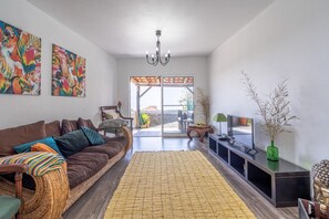 A living room with a brown sofa, colorful cushions, a woven rug, a TV on a black stand, and a green vase with branches. - Country House 'Serrão' with Private Terrace, Private Garden and Wi-Fi (Calheta)
