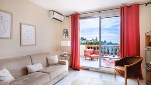 En stue med en beige sofa, et rundt sofabord og en rød lænestol. Væggen er prydet med indrammede kunstværker, et vindue med røde gardiner og udsigt til en udendørs patio med stol og bord.
