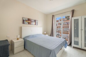 Quarto com cama, mesa de cabeceira, guarda-roupa e janela com vista para prédios.