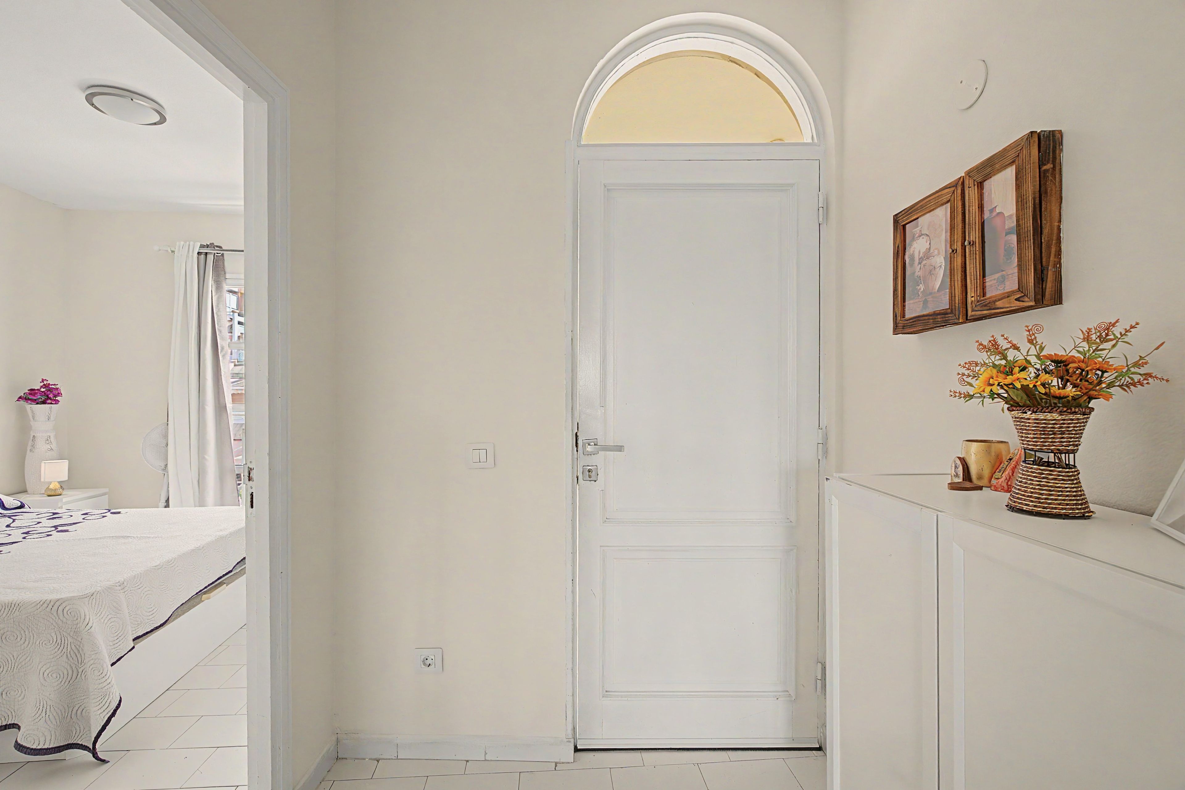 A hallway with a white door, a bed with a white bedspread, a white console table with a vase of flowers, and a wall-mounted picture frame.