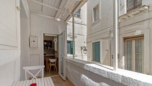 Un couloir étroit avec une table et des chaises blanches, une porte donnant sur une pièce et une fenêtre d’où l’on peut voir un immeuble.