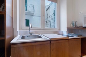 Una cocina con un lavamanos, armarios de madera, una ventana con vistas a edificios y una encimera con un hervidor y tazas.