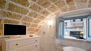 A room with a stone wall, a flat-screen TV on a dresser, a small table, and a window with a view of a building. - Apartment 'Pescatore' with Wi-Fi and Air Conditioning (Monopoli)
