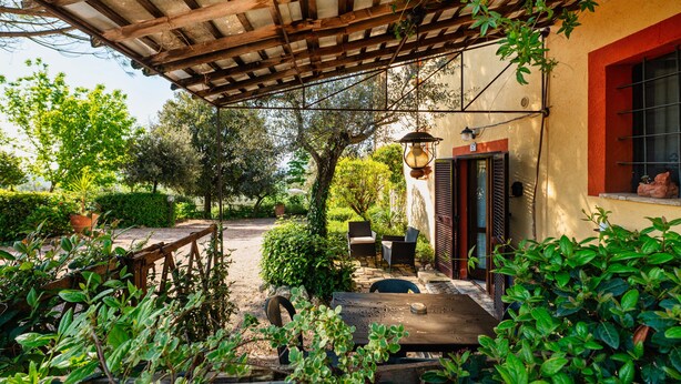 A covered patio with a table and chairs, surrounded by greenery and a building with a red window frame. - Apartment 'Merlot, 4' with Private Garden and Wi-Fi (Montefalco)