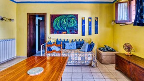 A room with a wooden dining table, a sofa with blue cushions, a colorful painting on the wall, and a window with curtains. - Apartment 'Merlot, 4' with Private Garden and Wi-Fi (Montefalco)