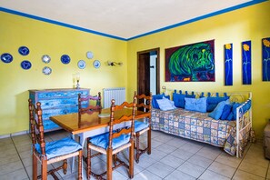 Een zithoek met een houten tafel en stoelen, een bank met blauwe kussens, en een schilderij aan de muur.