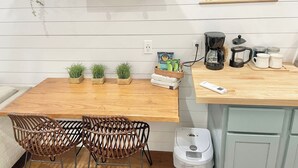 A kitchen with a wooden table, two wicker chairs, a coffee maker, a kettle, and a trash bin. - The Tiny with Big Time Vibes and Walkability (Austin)