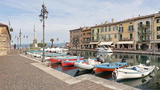 A waterfront with boats docked, buildings with balconies, and a paved walkway.
