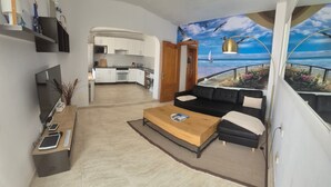 A modern living room with a sofa, coffee table, and a TV. There's a kitchen in the background and a balcony with a view of the sea. - Vacaciones 'La Orilla' with Private Terrace, Balcony, and Wi-Fi (Costa Calma)