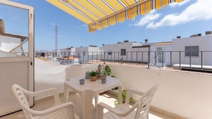 A balcony with a striped awning, white plastic chairs, a small table with a plant, and a view of neighboring buildings. - Vacaciones 'La Orilla' with Private Terrace, Balcony, and Wi-Fi (Costa Calma)