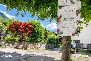 A sign for Hofladen Vendra Direct Sale with operating hours, a stone wall, a red flower bush, and a car parked in front. - Biobauernhof Toellerhof 'Room 5' with Mountain View, Shared Pool, and Wi-Fi (Algund)
