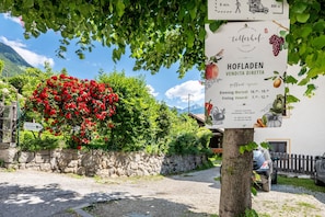 A sign for Hofladen Vendra Direct Sale with operating hours, a stone wall, a red flower bush, and a car parked in front.