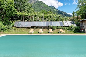 A pool with four sun loungers and solar panels in a lush, mountainous setting.
