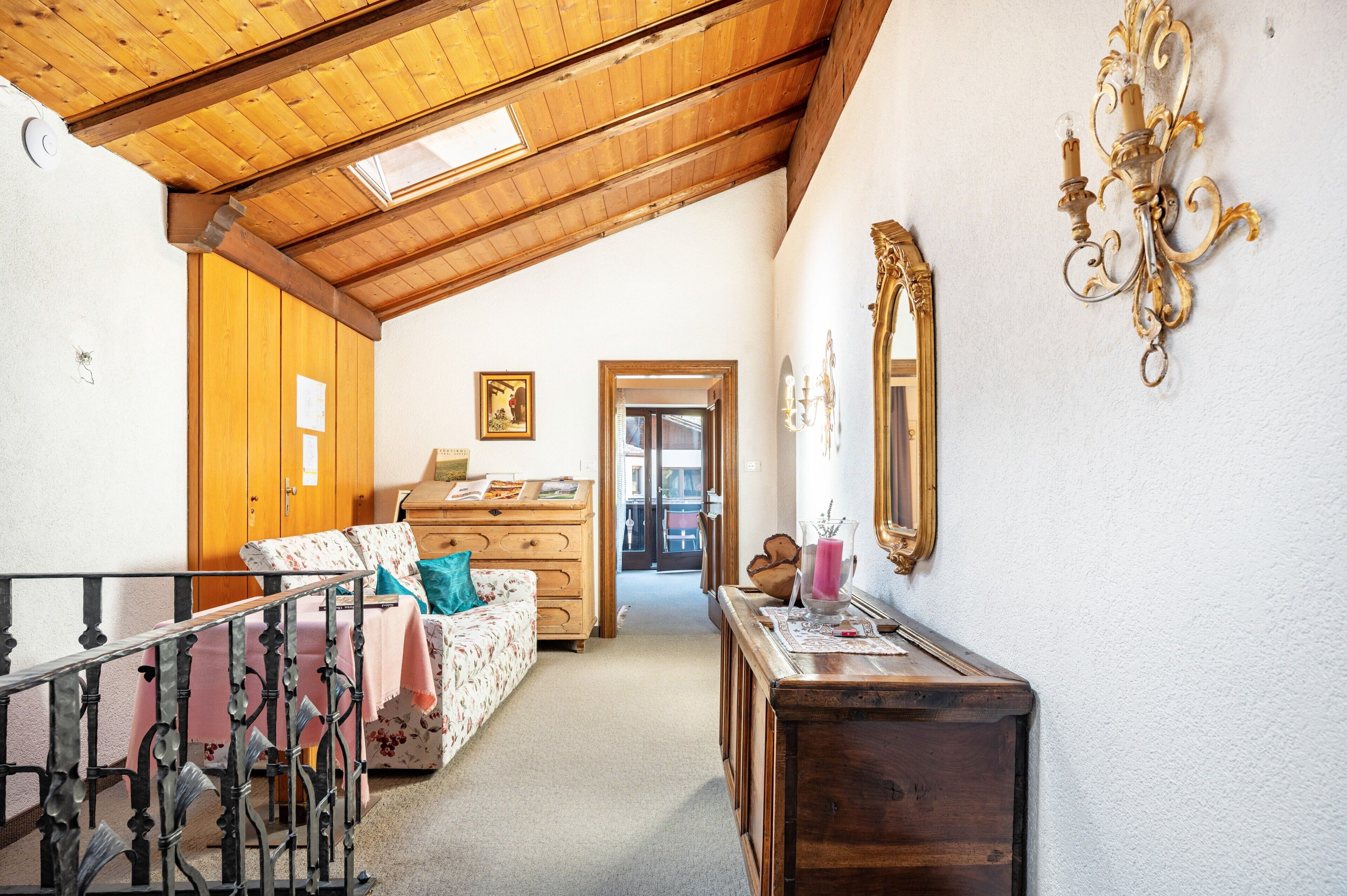 A room with a wooden ceiling, a bed with floral bedding, a dresser, a mirror, and a door leading to another room.