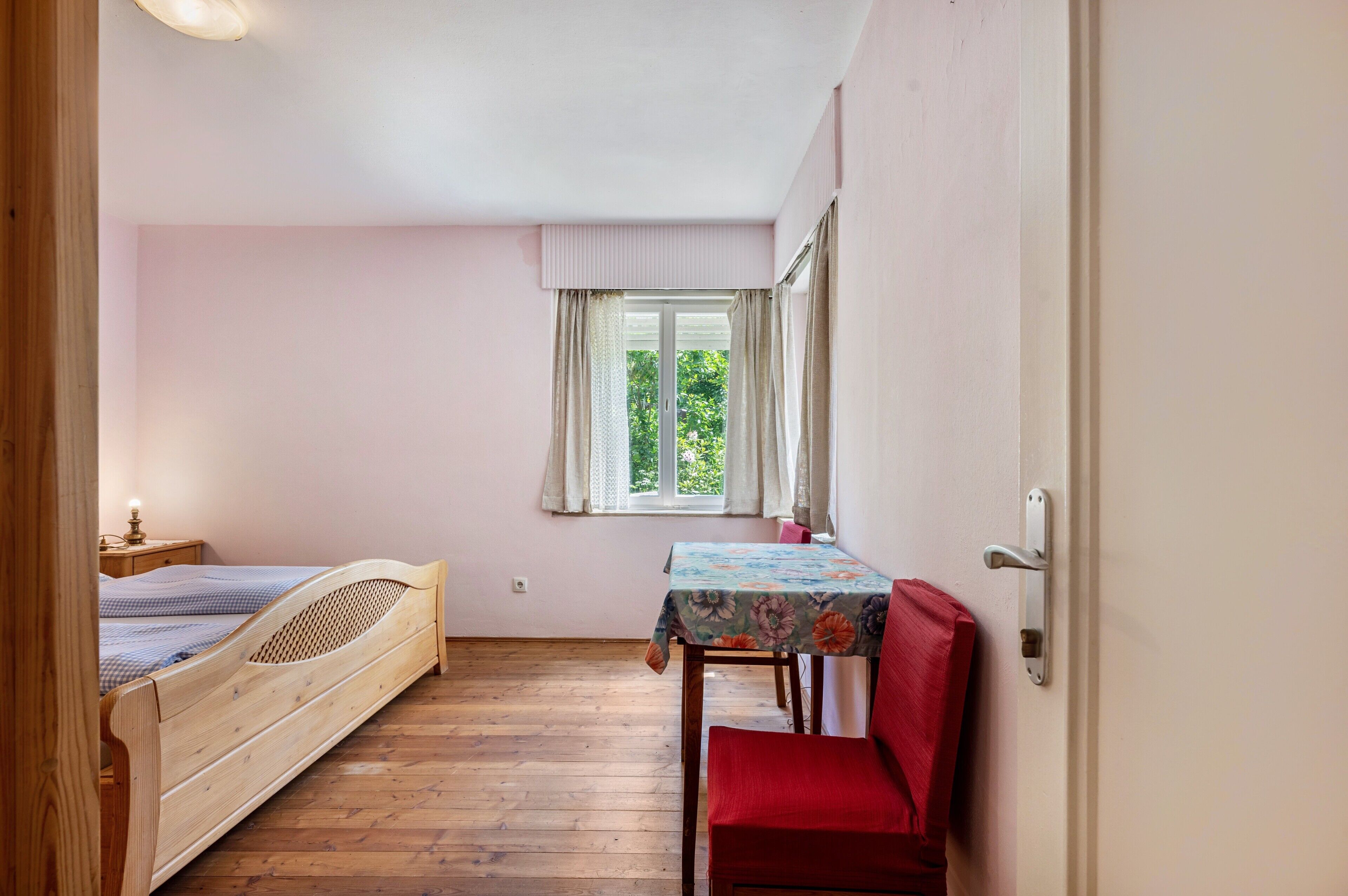 Une chambre à coucher avec un lit en bois, une petite table recouverte d’une nappe à motifs floraux, une chaise rouge et une fenêtre avec des rideaux.