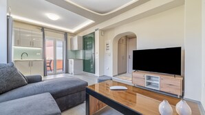 A modern living room with a sofa, a coffee table, a flat-screen TV, and a kitchen area visible through a glass door. - Luxury Stay Apartment with Mountain View, Wi-Fi, and Air Conditioning (Kerkira)