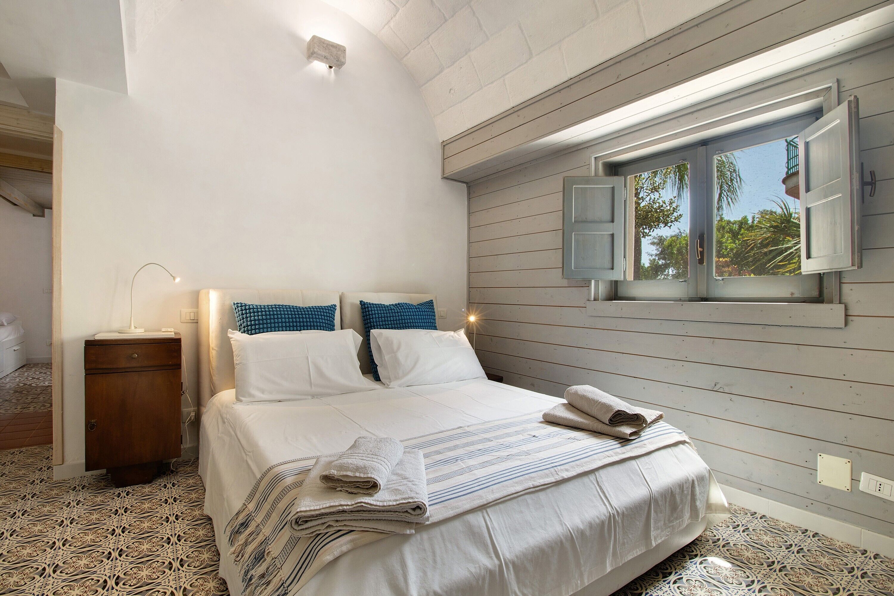A bedroom with a bed, pillows, a nightstand, a window with shutters, and a patterned floor.