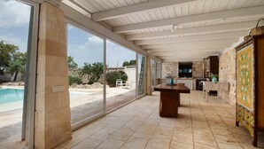 Une pièce spacieuse dotée d’une grande porte-fenêtre coulissante donnant sur une piscine, d’une table en bois et d’un coin cuisine aux murs en pierre.