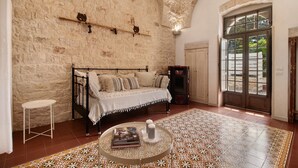 Une chambre avec un mur en pierre, un lit recouvert d’une courtepointe blanche, une table basse ronde ornée de livres et d’une bougie, et un tapis à motifs.