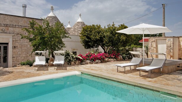 Un espace piscine agrémenté de chaises longues et d’un parasol, entouré de bâtiments en pierre et de plantes en fleurs.