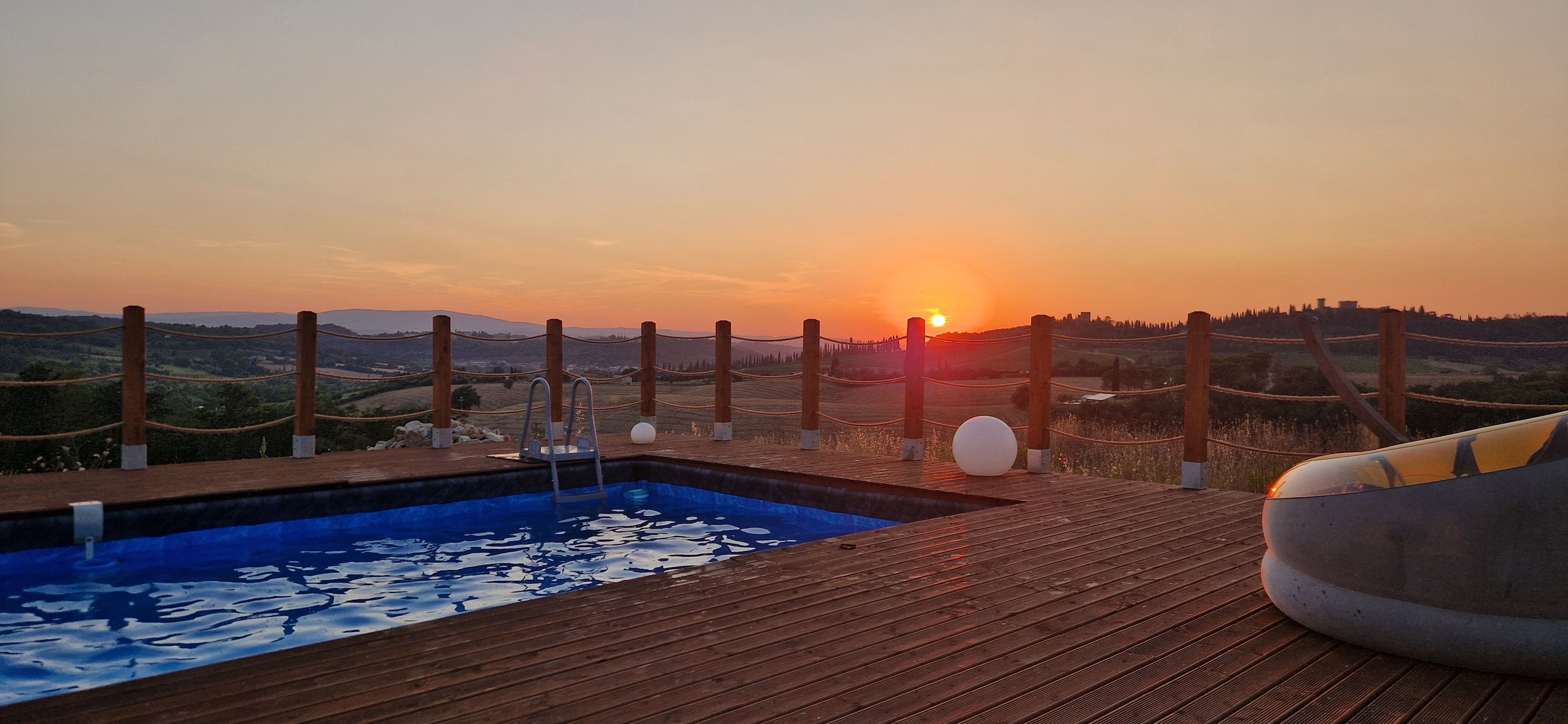 Una terraza de madera con piscina, una tumbona y vistas a la puesta de sol sobre un paisaje montañoso.