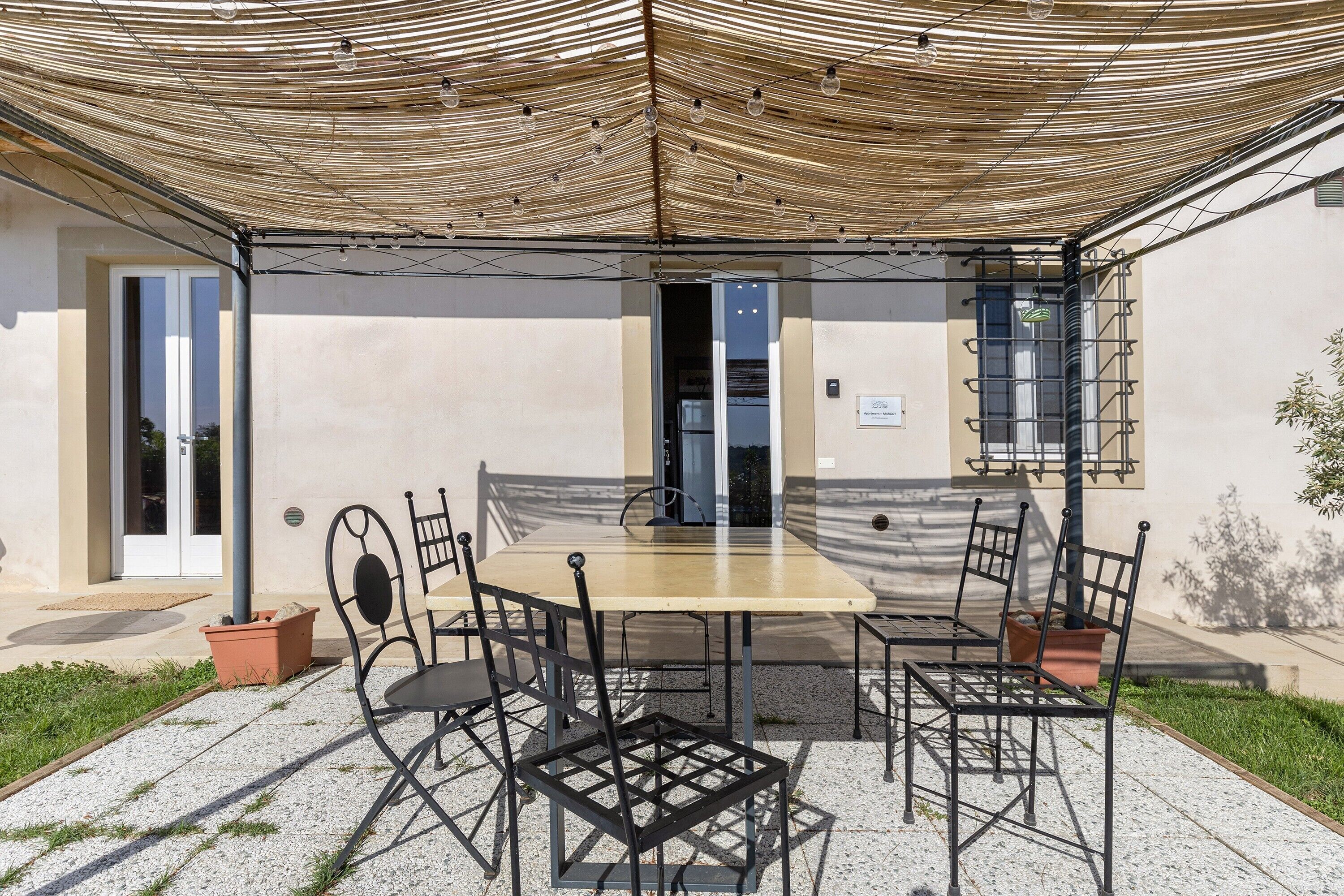Un patio exterior con mesa y sillas, una pérgola y la entrada a un edificio.