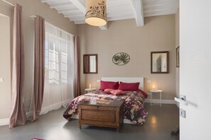 Une chambre à coucher avec un lit, une table de chevet, une lampe, un miroir et une fenêtre avec des rideaux.