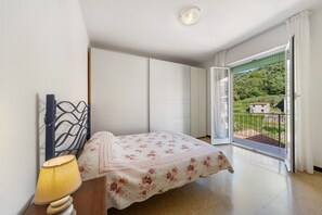Een slaapkamer met een bed, een lamp, een raam met uitzicht op groen en een balkon.