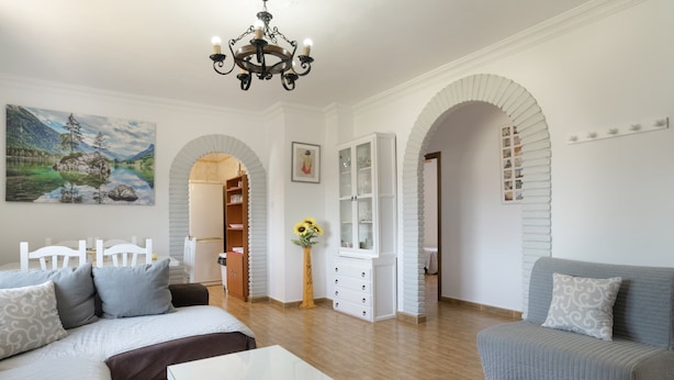 A living room with a sofa, a glass coffee table, a chandelier, a painting, and an arched doorway.