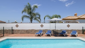 A swimming pool with lounge chairs and umbrellas, palm trees, and a white wall with a stone base.