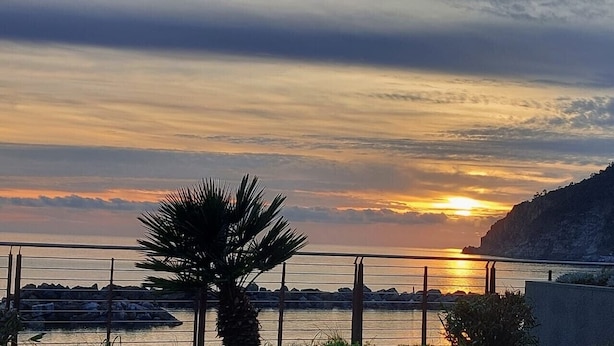 A sunset over a coastal landscape with a palm tree in the foreground and a rocky cliff in the background. - Apartment for 5 Guests Steps from the Sea (Moneglia)