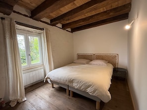 A bedroom with a wooden bed, a window with curtains, a radiator, and a small bedside table.