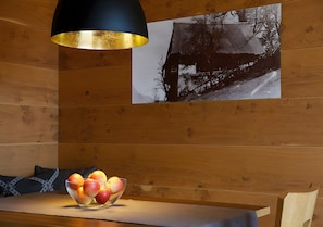 Un intérieur en bois avec une table, un saladier de fruits, un luminaire suspendu noir et doré et une photographie en noir et blanc accrochée au mur.