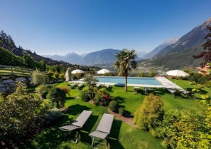 Zona de piscina con sillas de descanso y sombrillas, rodeada de follaje cuidado y montañas al fondo.
