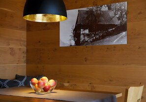 Un interior de madera con una mesa, un cuenco de fruta, una lámpara colgante negra y dorada, y una fotografía blanco y negro en la pared.