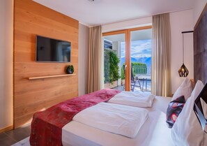 A hotel room with a bed, a TV mounted on a wooden wall, a balcony with a view, and a dining area visible through the glass doors. - Apartment 'Sonnenblume' with Mountain View, Shared Pool and Wi-Fi (Schenna)