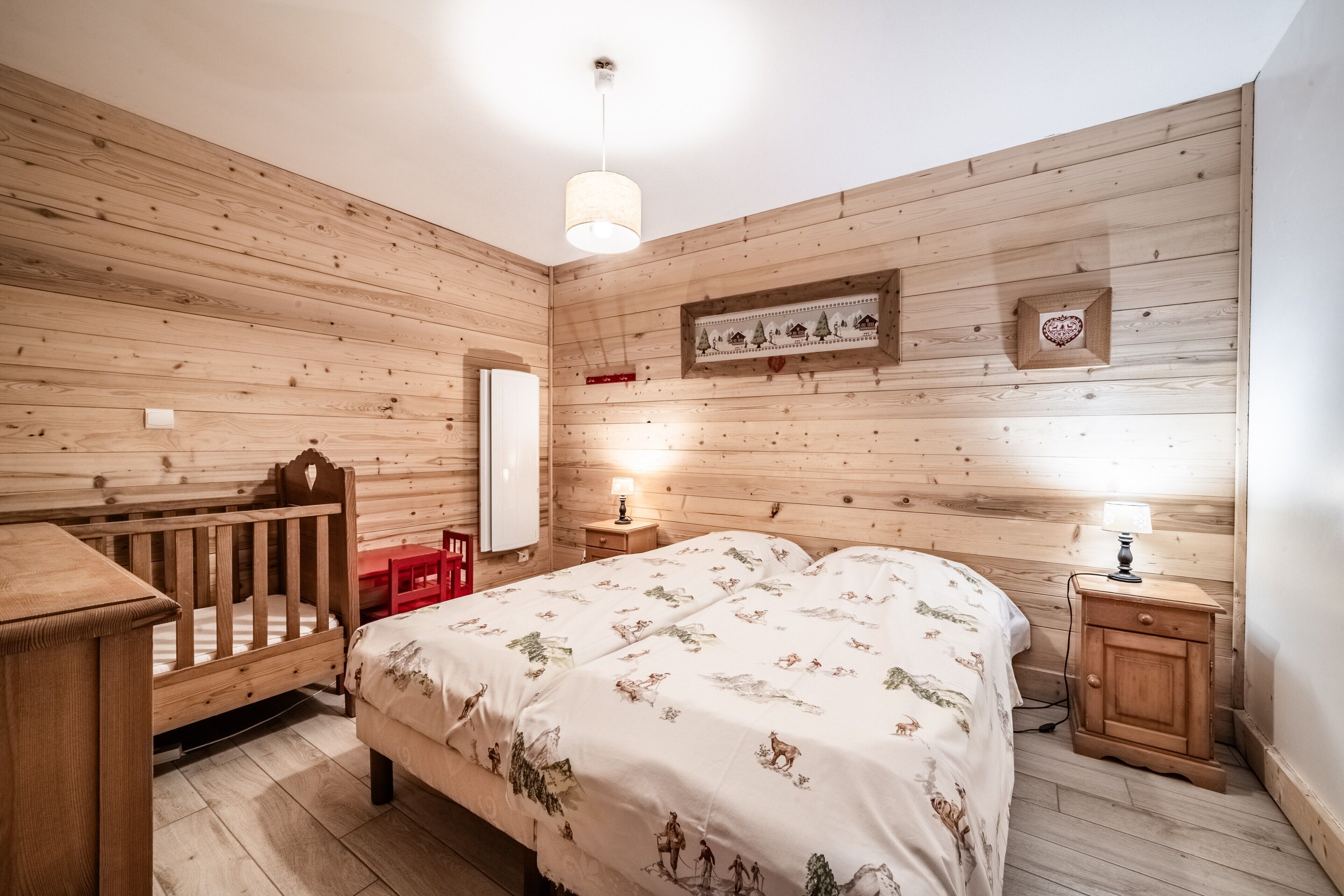 Habitación con paneles de madera, una cama, una cuna, una mesita de noche, una lámpara y un espejo.