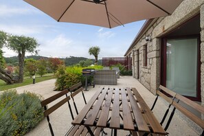 Une terrasse extérieure avec une table et des bancs en bois, un grand parasol et un bâtiment en pierre doté d’une porte en verre.