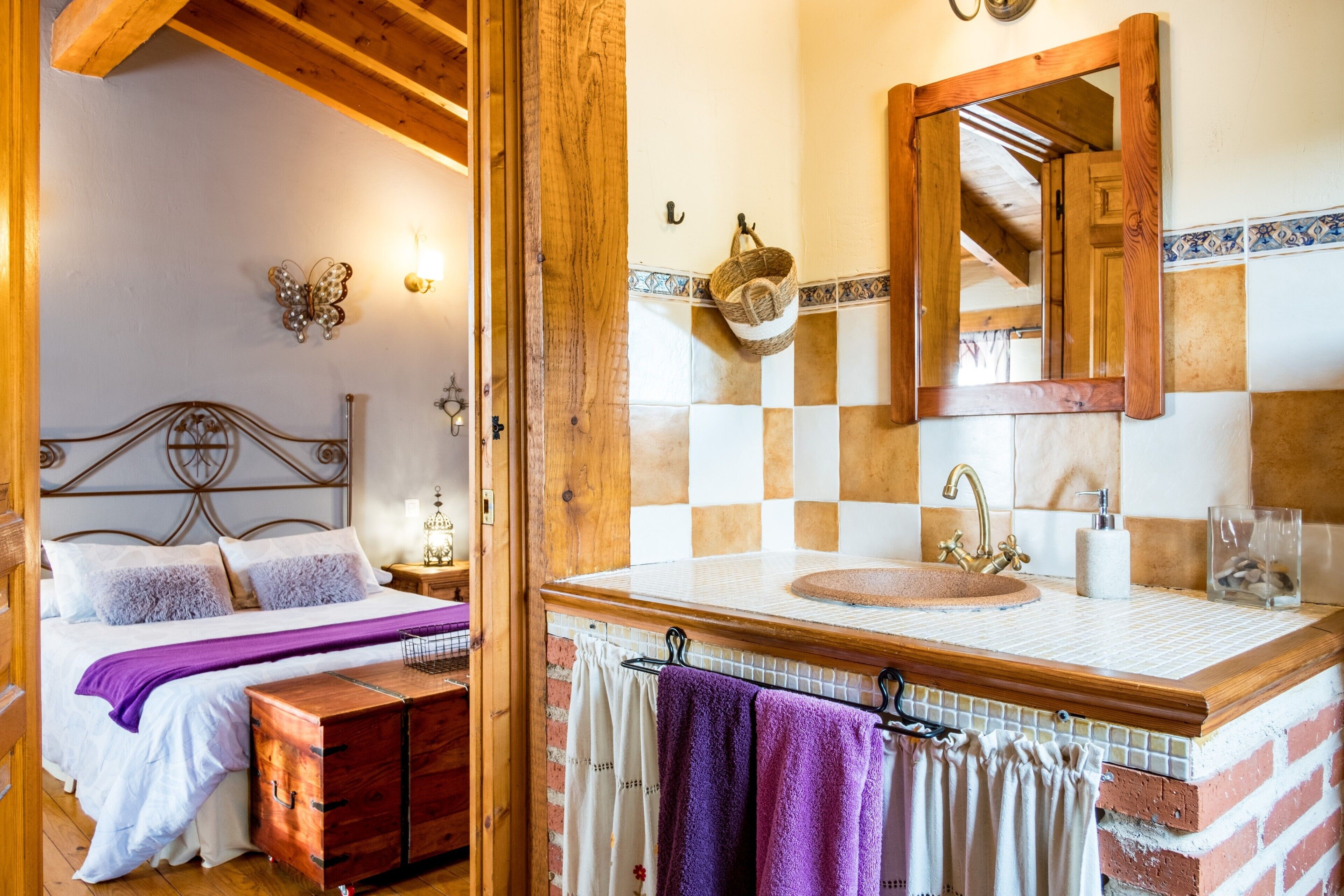 Een slaapkamer met een bed, een houten nachtkastje, een dressoir, en een badkamer met een wastafel, spiegel en betegelde achterwand.