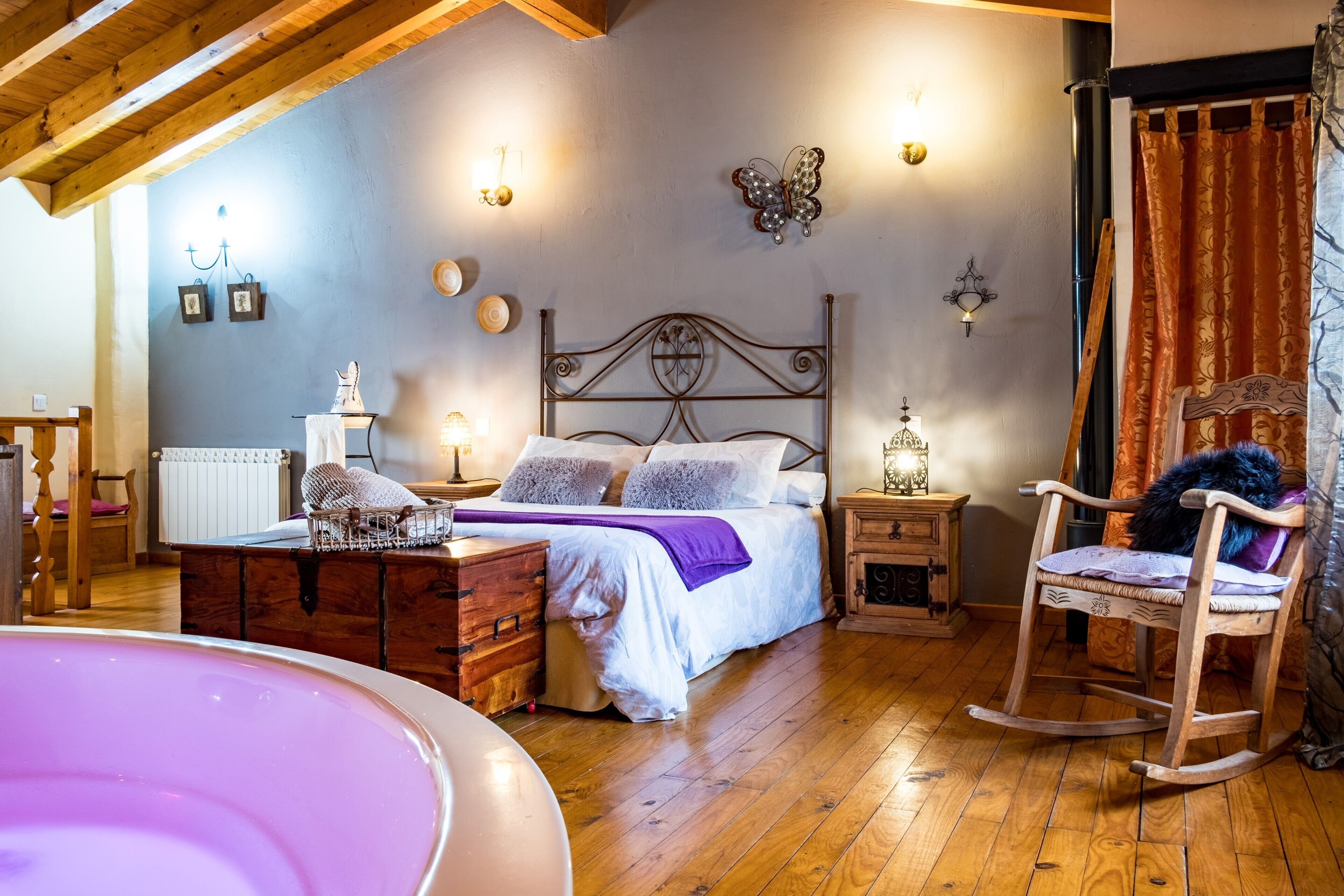 A bedroom with a wooden ceiling, a bed with white bedding, a wooden rocking chair, a dresser, and a nightstand with a lamp.