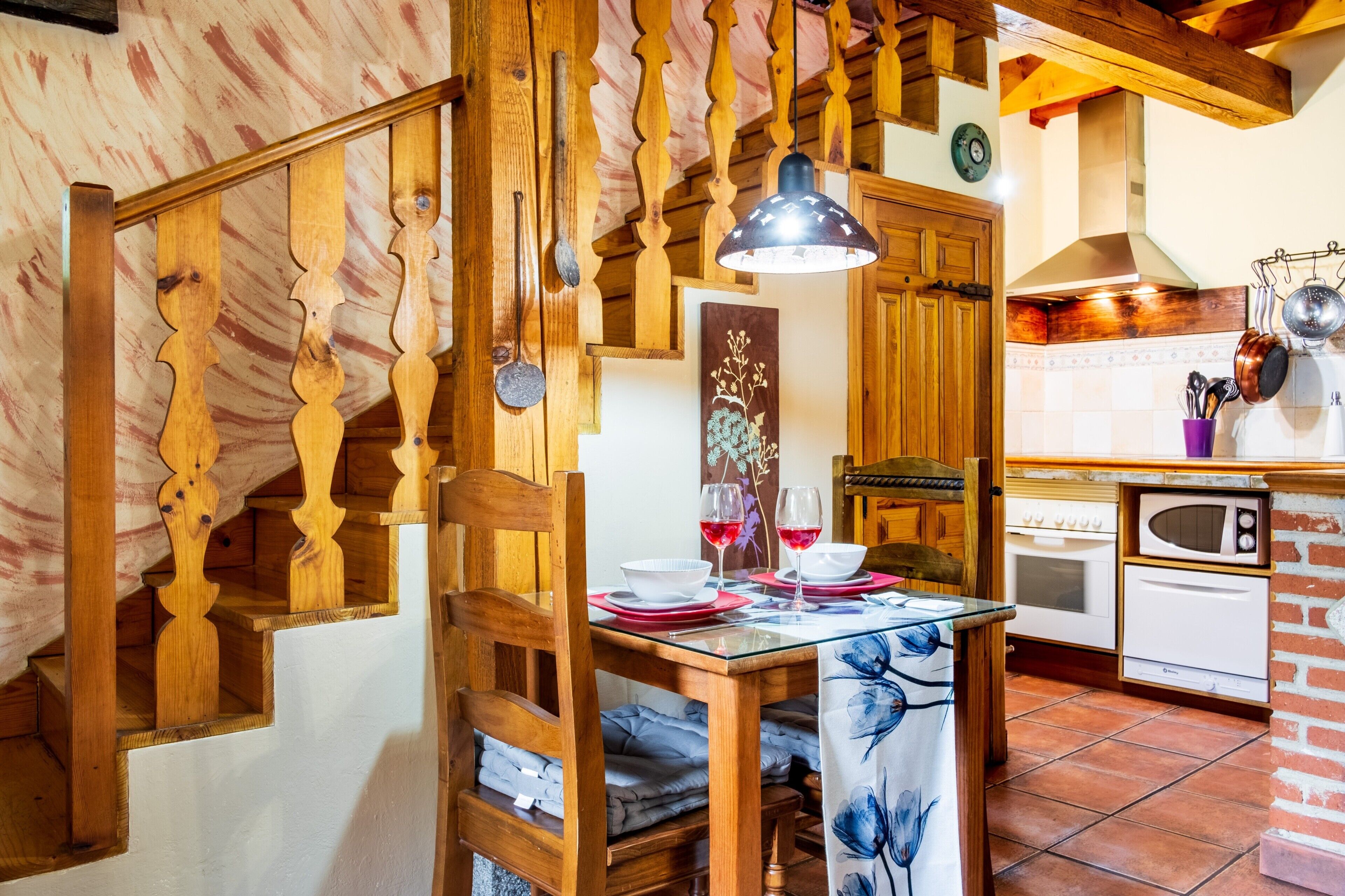 Een houten trap met een glazen tafel gedekt voor twee personen, een hanglamp en een keuken met witte apparaten.