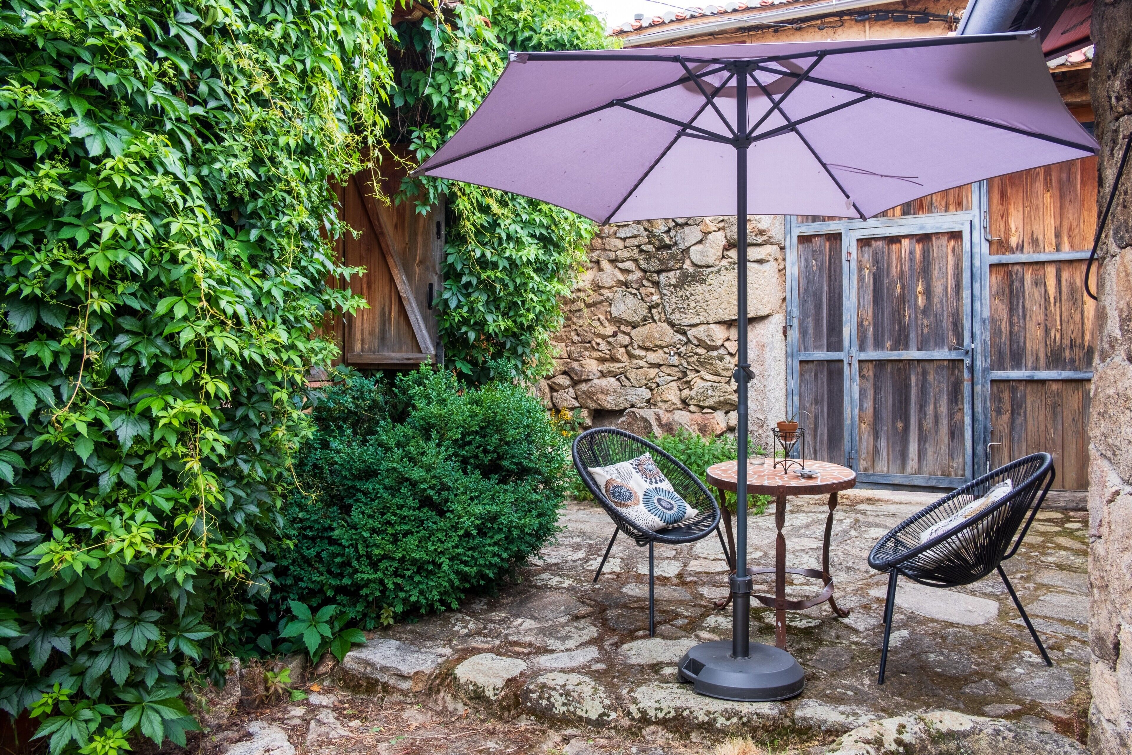 A patio with a table and chairs, surrounded by greenery and stone walls.