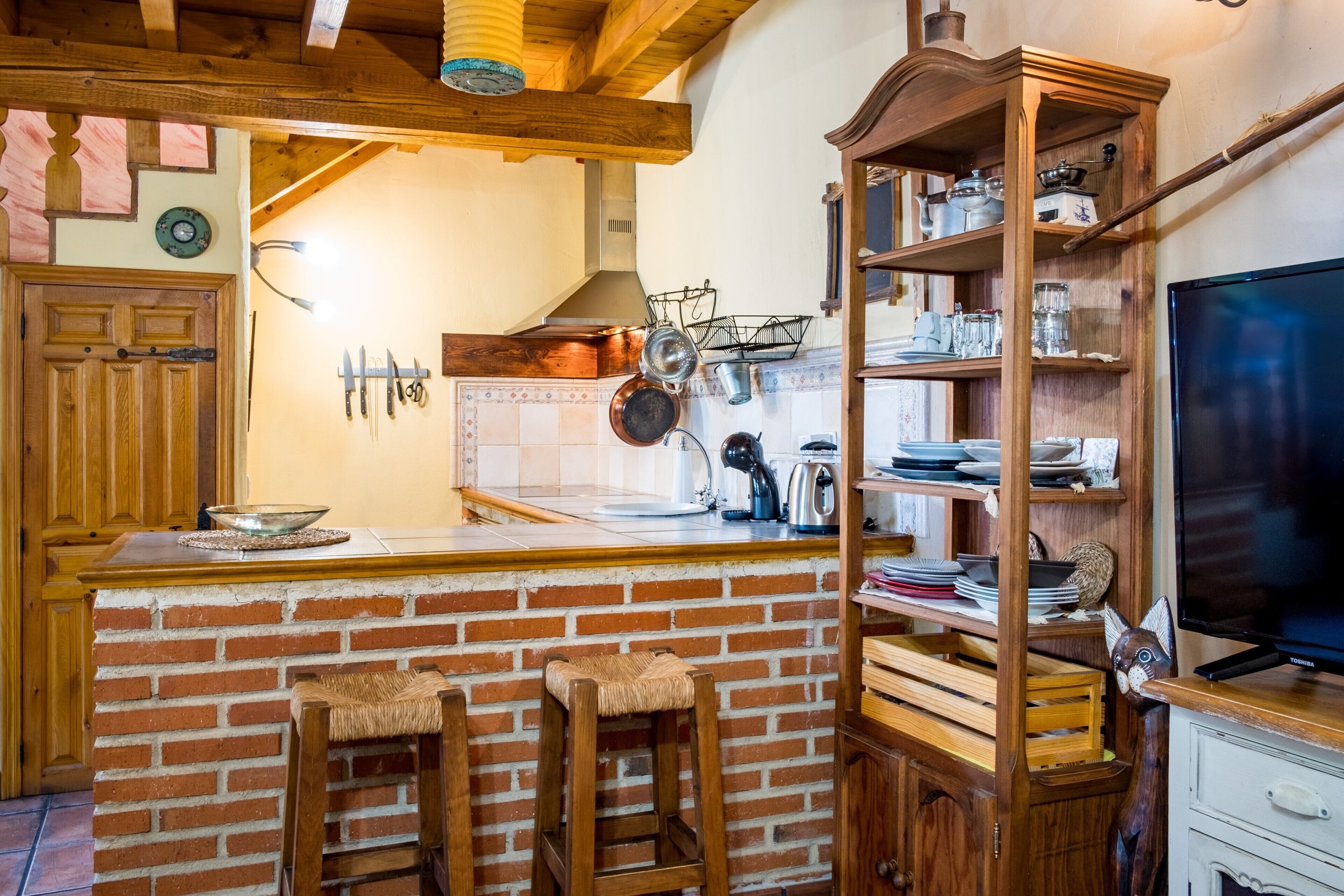 Une cuisine avec un îlot central en briques, des tabourets en bois, un téléviseur et une étagère contenant des ustensiles de cuisine.