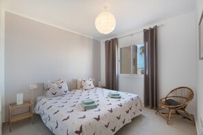 A bedroom with a bed, a bedside table, a chair, a window with curtains, and a hanging light fixture.