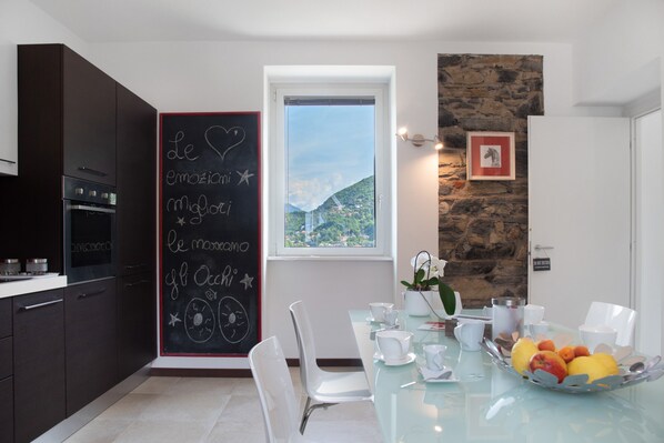 Une cuisine moderne avec des meubles de couleur sombre, un tableau noir avec des inscriptions, un coin repas avec une table en verre, des chaises et un panier de fruits.