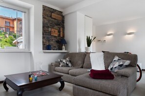 Un salon comprenant un canapé, une table basse, un mur en pierre et une fenêtre donnant sur des immeubles.