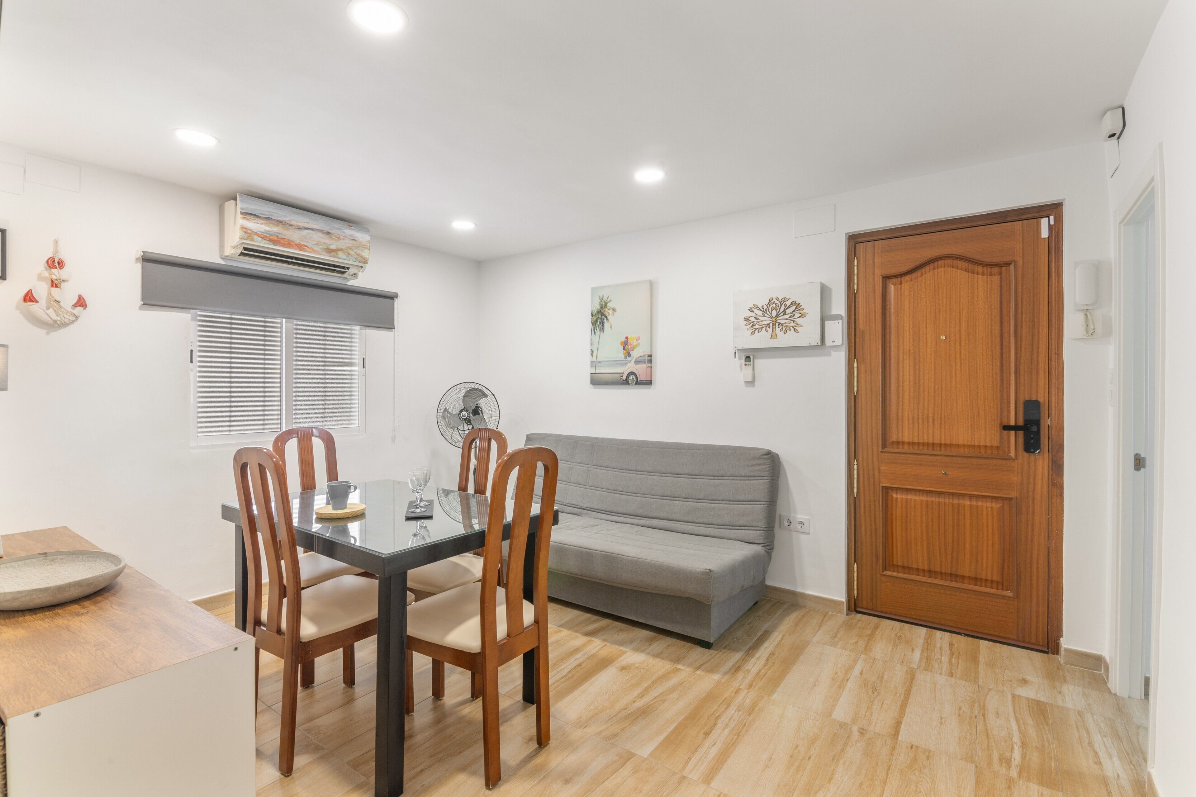 A dining area with a table, chairs, a fan, a grey sofa, and a wooden door.