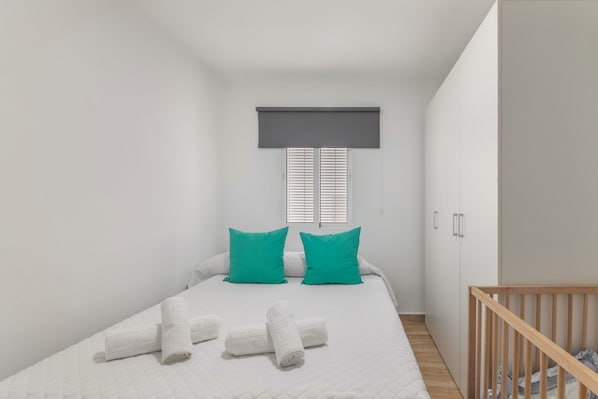 Un dormitorio con cama, almohadas, una ventana con persianas, un armario y una cuna.